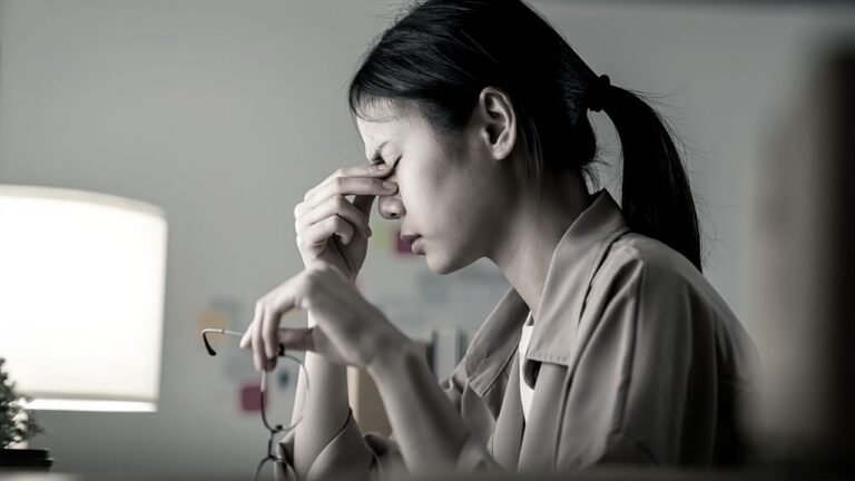 A woman removing her glasses to rub the tension from her eyes. The stresses in the workplace can take it's toll on all.