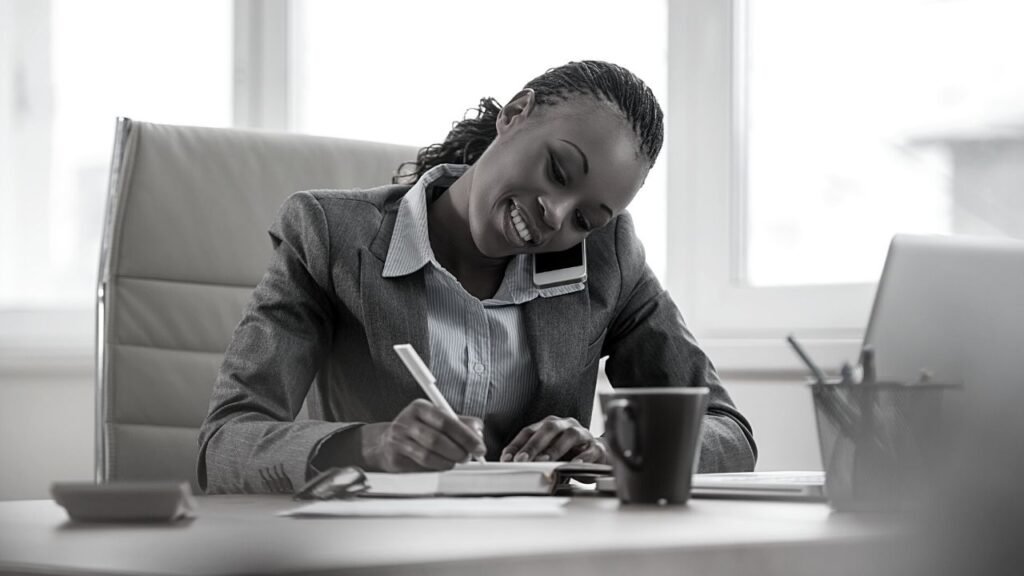 Business woman in a suit seated at a desk writing notes whilst on the phone talking with The Ethical Strategist about the new love for the business with HouseRules