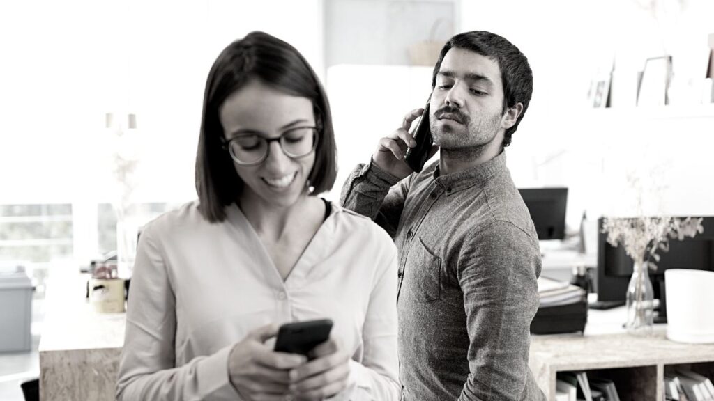 A man peeping over the shoulder of a woman as she catches up on the latest Ethical Insights on her phone. When considering remote working practices, you should consider the security of your business